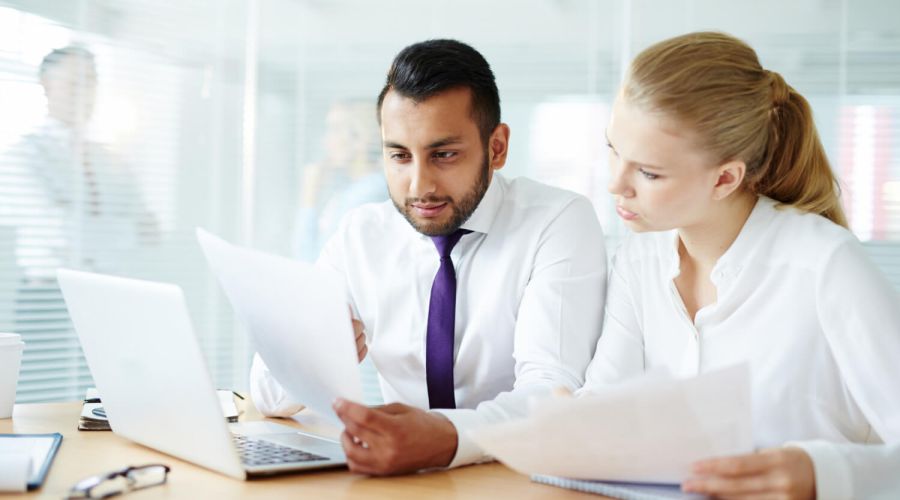 Two office colleagues reviewing documents together at a desk beside a laptop.