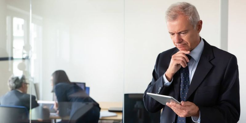 Older man in a suit holds a tablet, pondering thoughtfully. In the background, two colleagues converse in a glass-walled office, creating a focused work atmosphere.