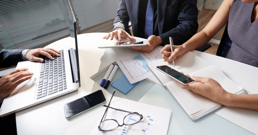 Three professionals gathered around a meeting table, collaborating with a laptop, tablet, smartphone, notebook, printed charts and documents during a business planning session in a modern office.