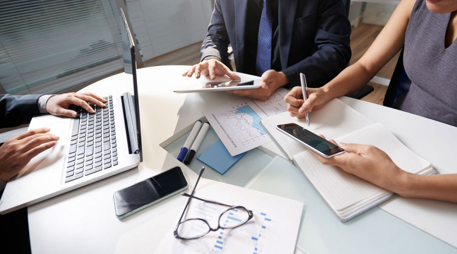 Three professionals gathered around a meeting table, collaborating with a laptop, tablet, smartphone, notebook, printed charts and documents during a business planning session in a modern office.