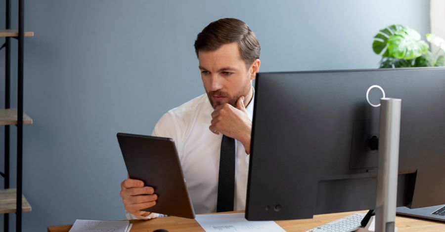 Business professional reviewing information on a tablet while working at a desktop computer in a modern office.