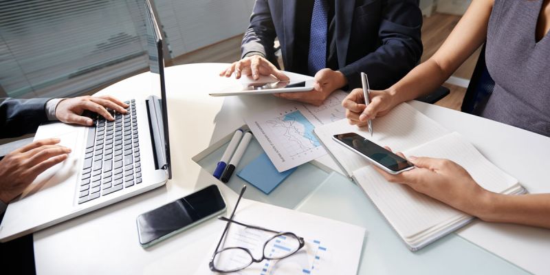 Three professionals gathered around a meeting table, collaborating with a laptop, tablet, smartphone, notebook, printed charts and documents during a business planning session in a modern office.
