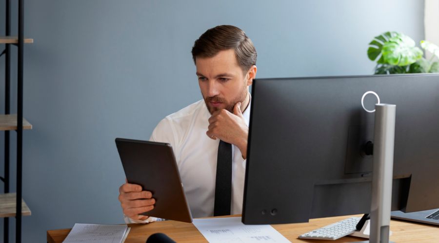 Business professional reviewing information on a tablet while working at a desktop computer in a modern office.