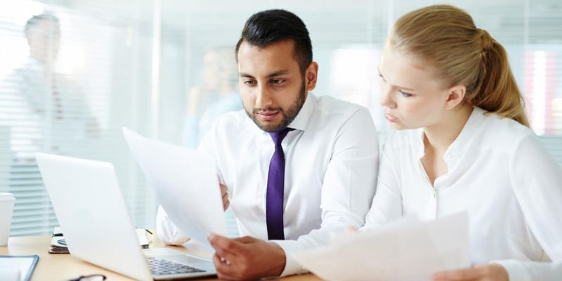 Two office colleagues reviewing documents together at a desk beside a laptop.